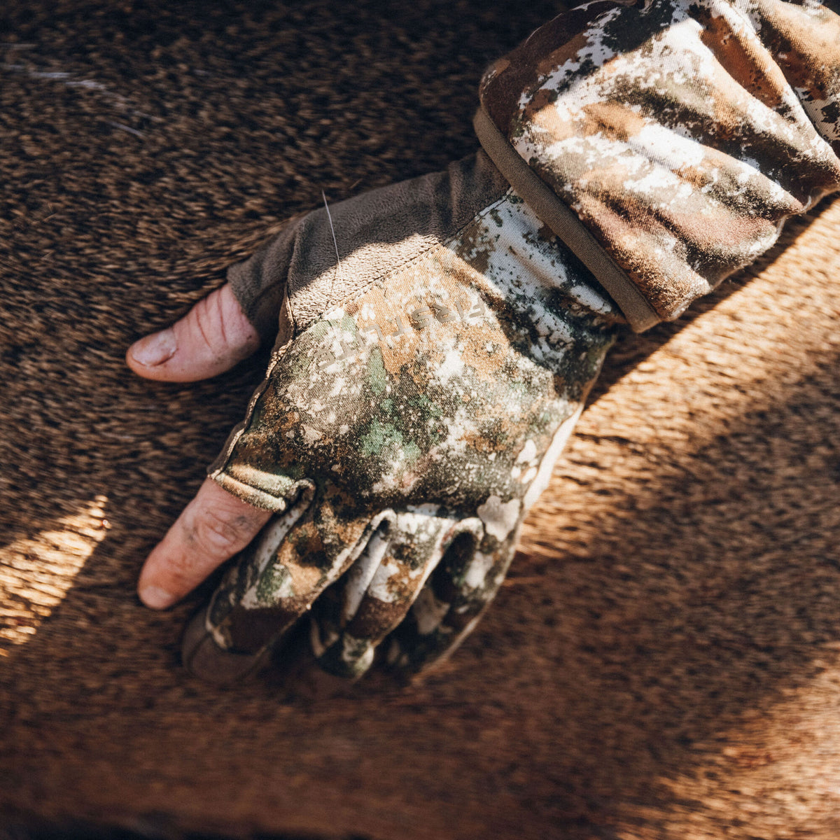 A brown, green, and white camouflage fingerless glove featuring a 'FIRSTLITE' logo is worn on a hand resting on brown fur.