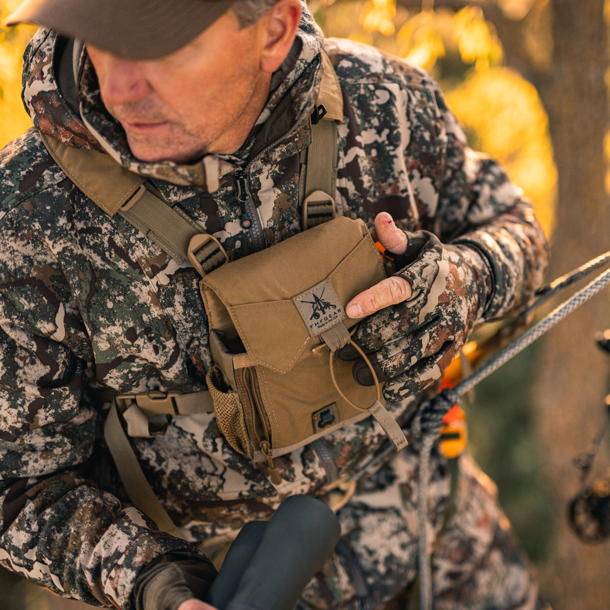 Tan FHFGEAR binocular chest harness with drawstring closure and mesh side pocket, worn by a person in camouflage.