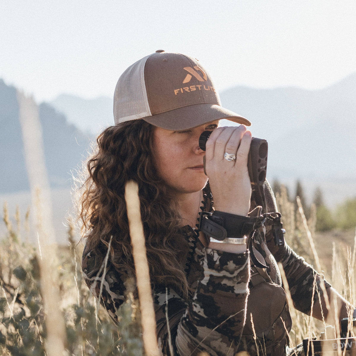 A woman in a brown and tan First Lite trucker hat and camouflage clothing looks through a dark brown rangefinder in a field of tall grass.