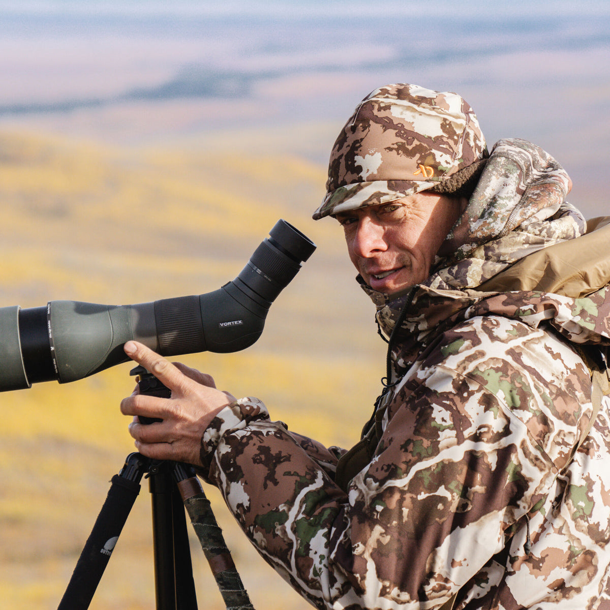 A person in brown and green camouflage operates a dark green Vortex spotting scope mounted on a tripod.