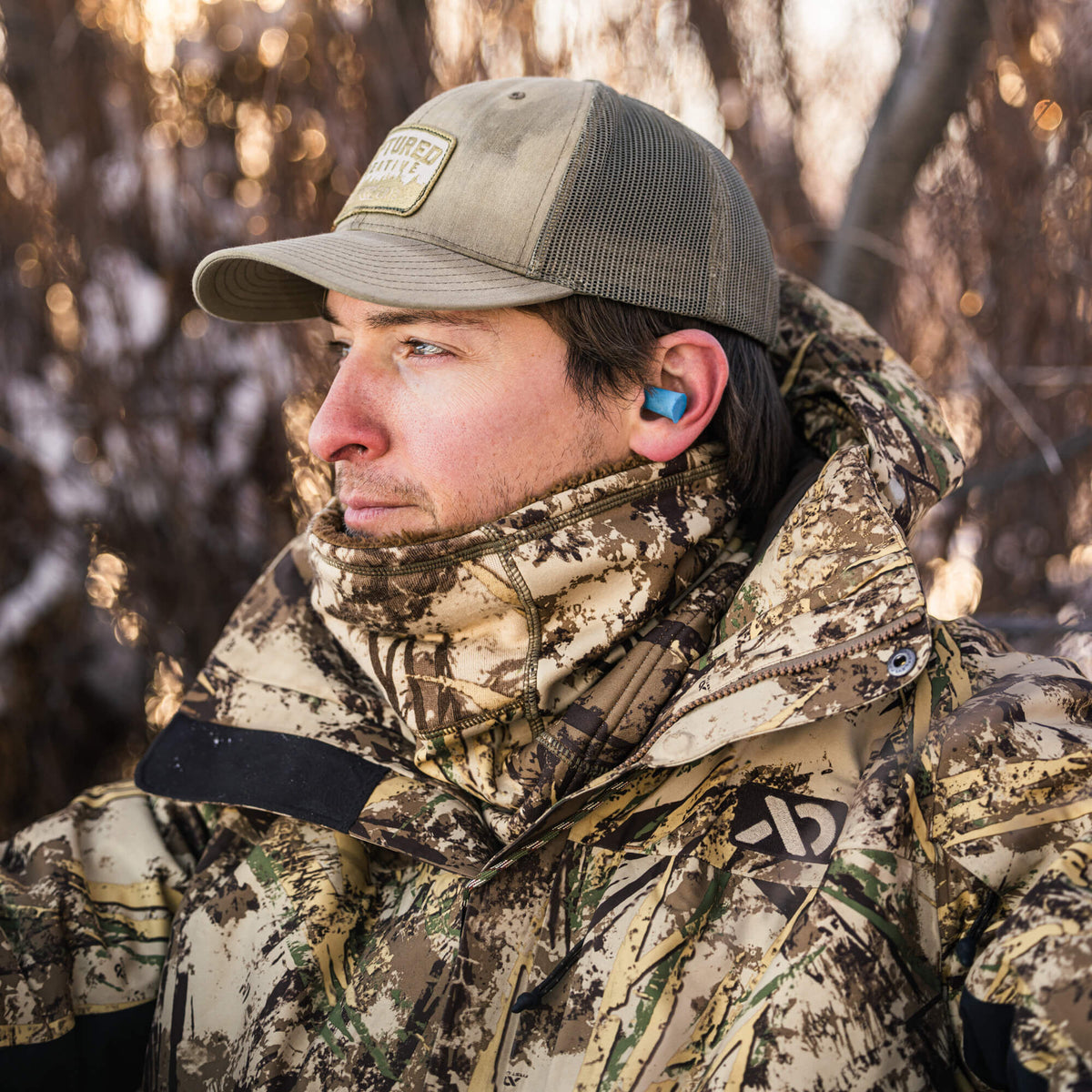 Man wearing bright blue earplugs in his right ear, a tan trucker cap, and a camouflage jacket.
