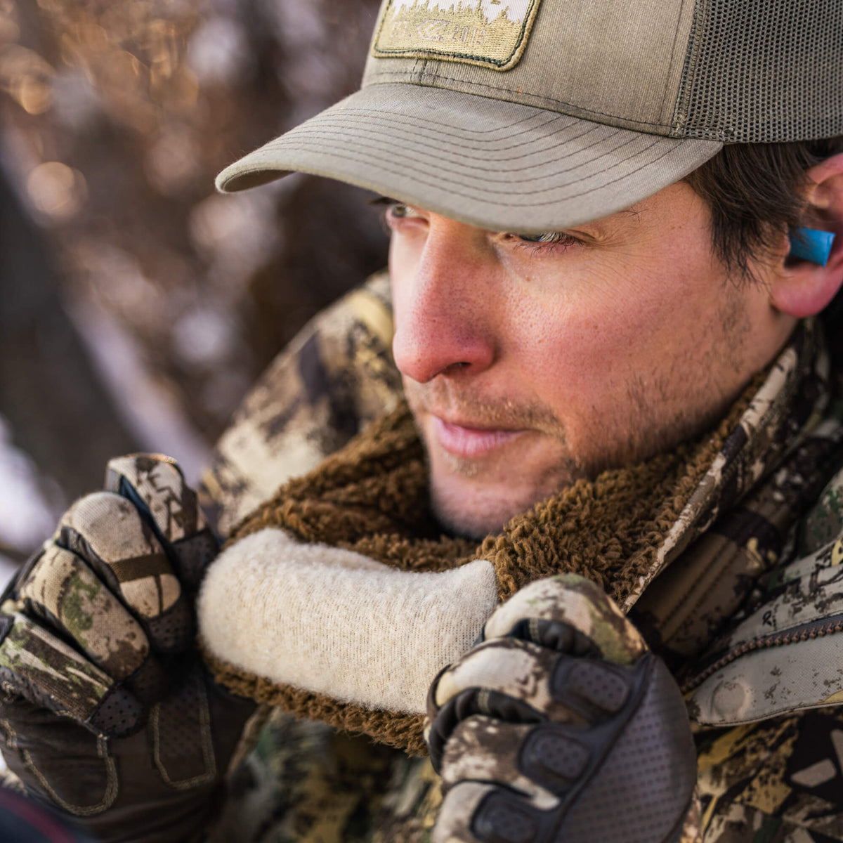 A man wearing an olive green cap with a patch, camouflage jacket, and gloves adjusts a cream knitted neck gaiter with brown fleece trim, showing blue earplugs.