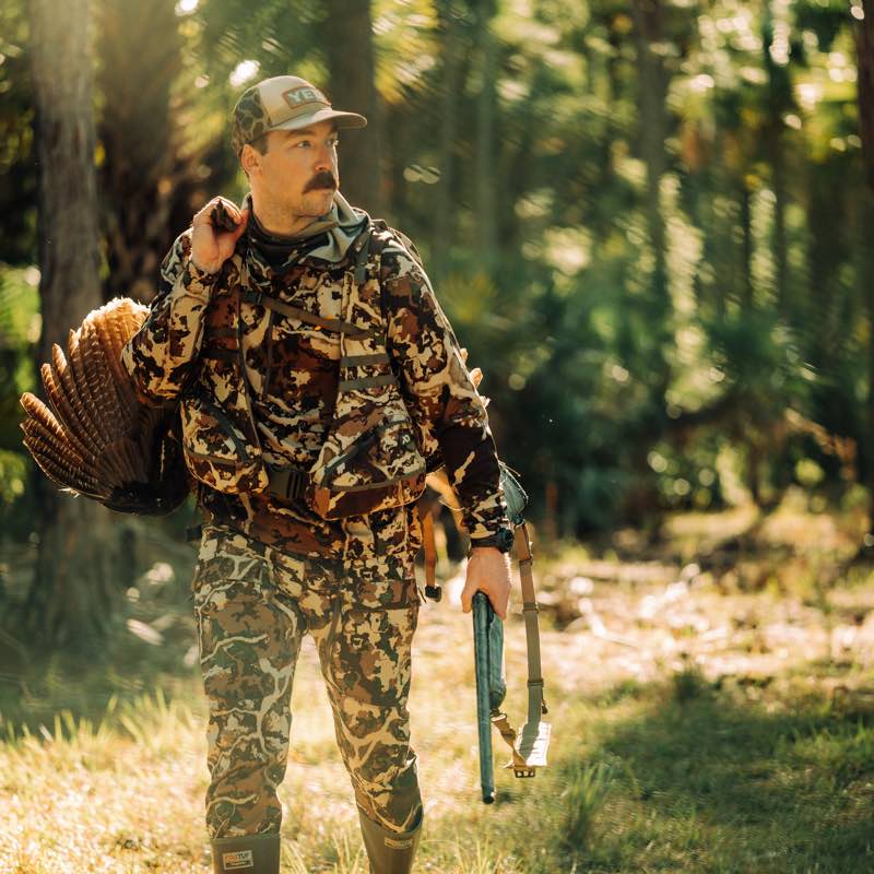 Man in brown/green camo hunting gear, YETI hat, carrying turkey and shotgun.
