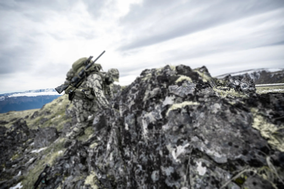 Camo person with olive pack, dark rifle with scope climbs mossy rocks.