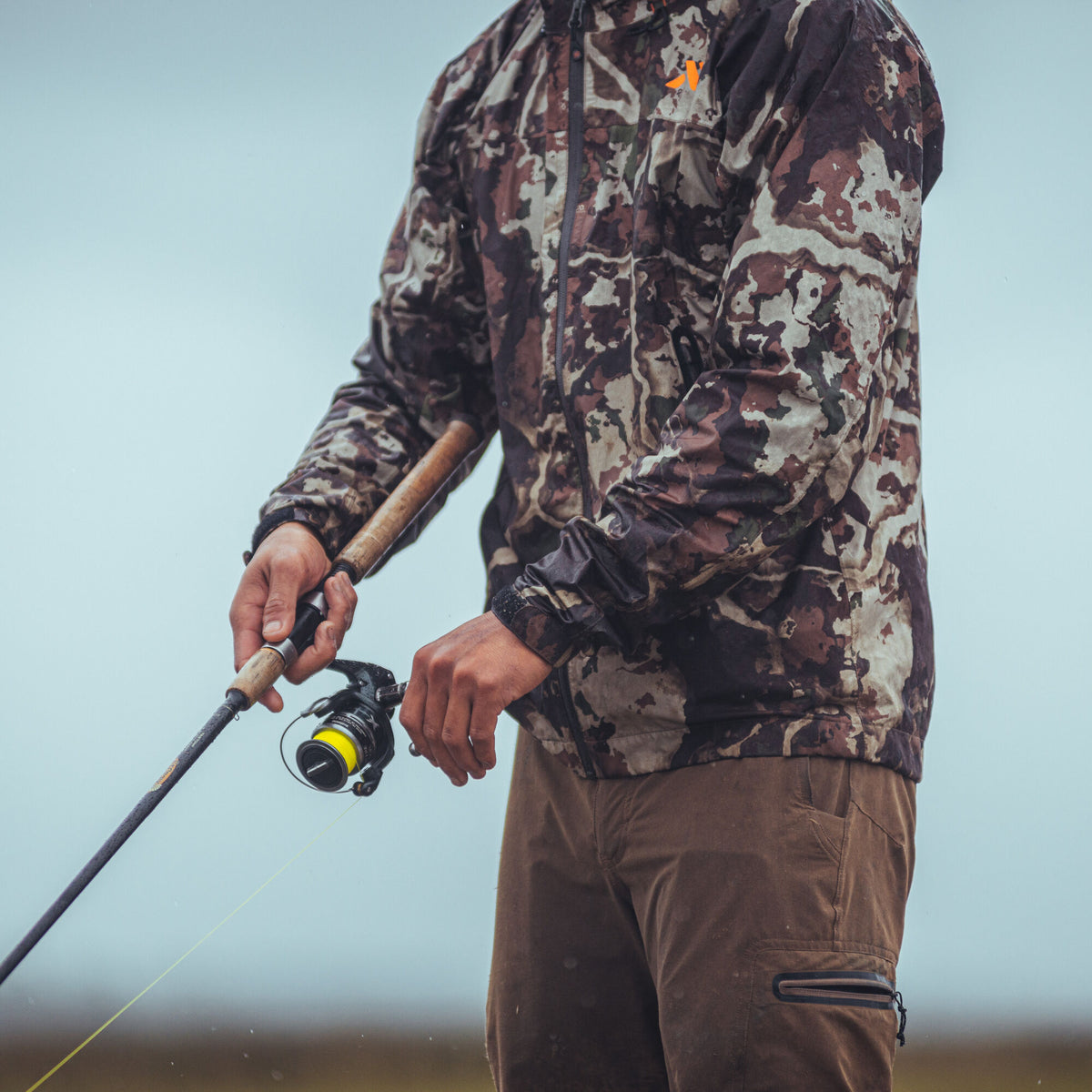 A person wears a brown and tan camouflage outdoor jacket with an orange chest logo and brown pants with a zippered side pocket, holding a fishing rod with bright yellow line.