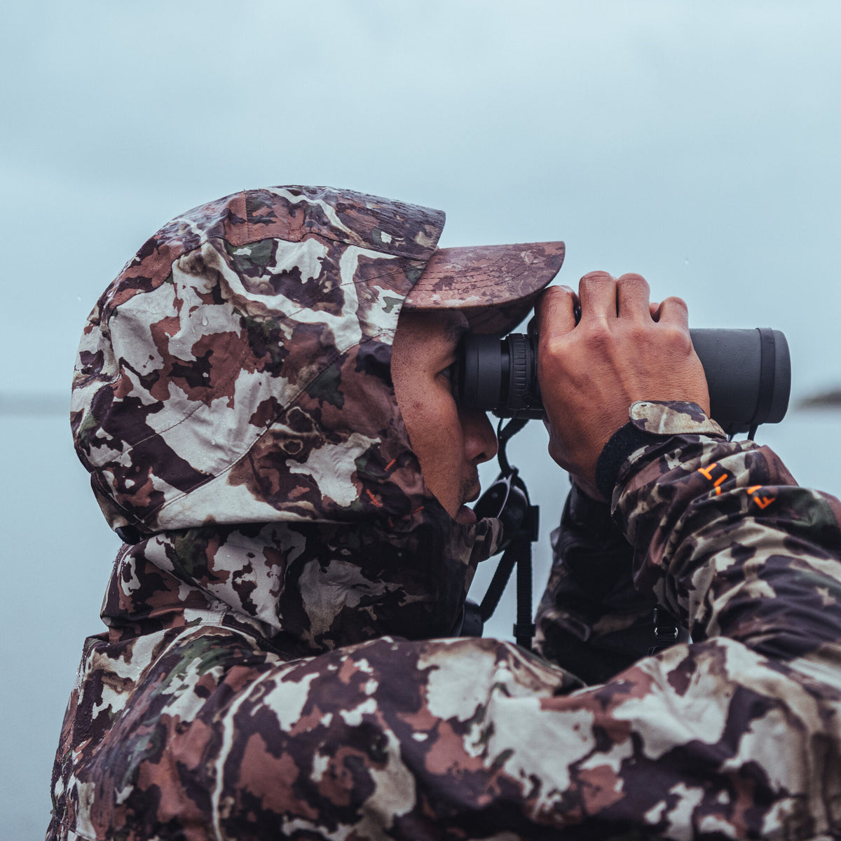 Hooded earth-toned camouflage rain jacket with visible water droplets and an orange First Lite logo on the sleeve.