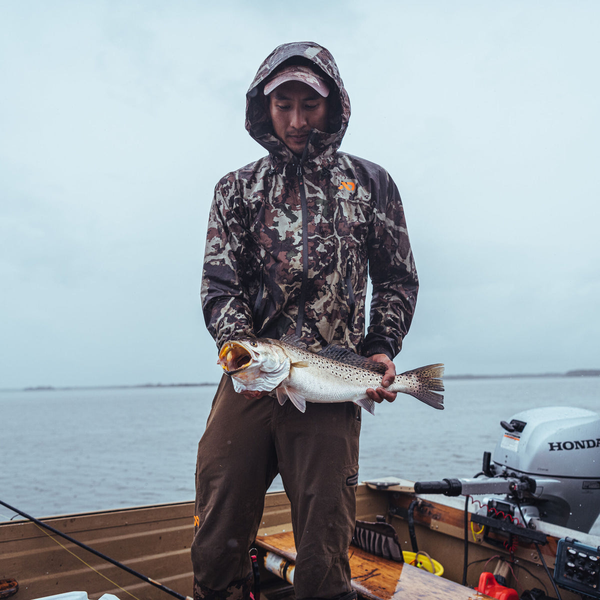 A man wearing a brown camouflage hooded rain jacket with an orange chest logo holds a speckled trout with its mouth open on a boat.