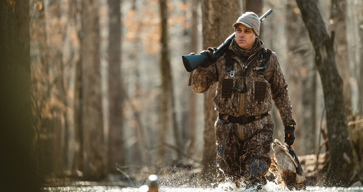 Hunter in camo waders carries shotgun and duck, splashing through water.