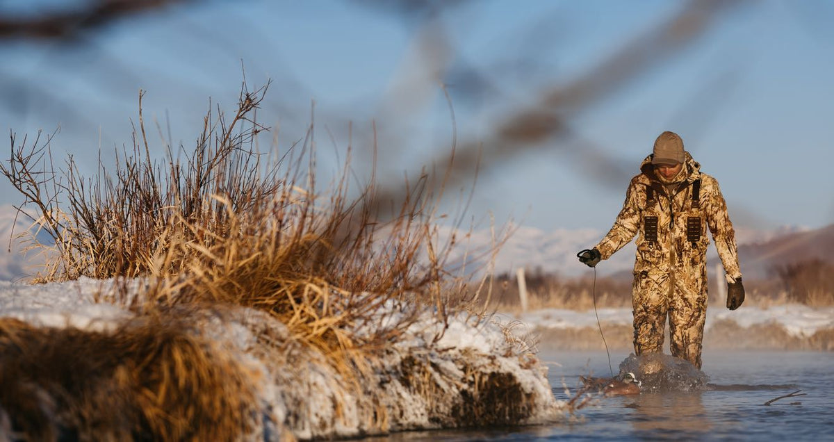 Tan camo hunting suit with tactical webbing, worn by hunter wading in water with a duck decoy.