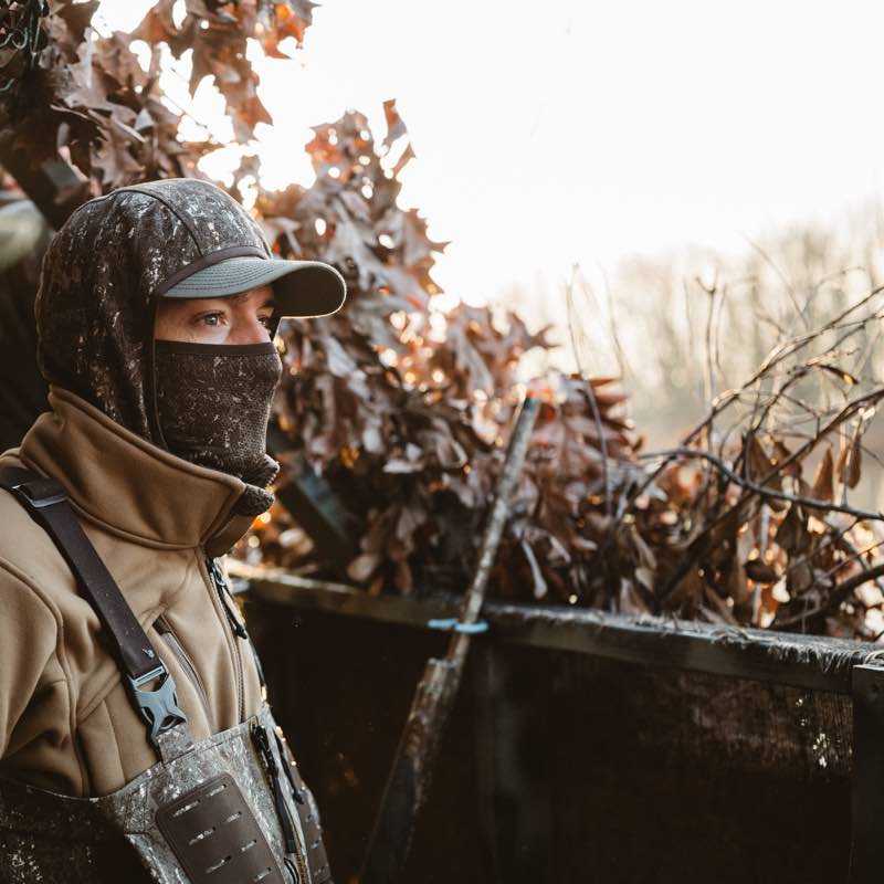 Hunter wears tan jacket, camo hooded balaclava, and bibs in a hunting blind.