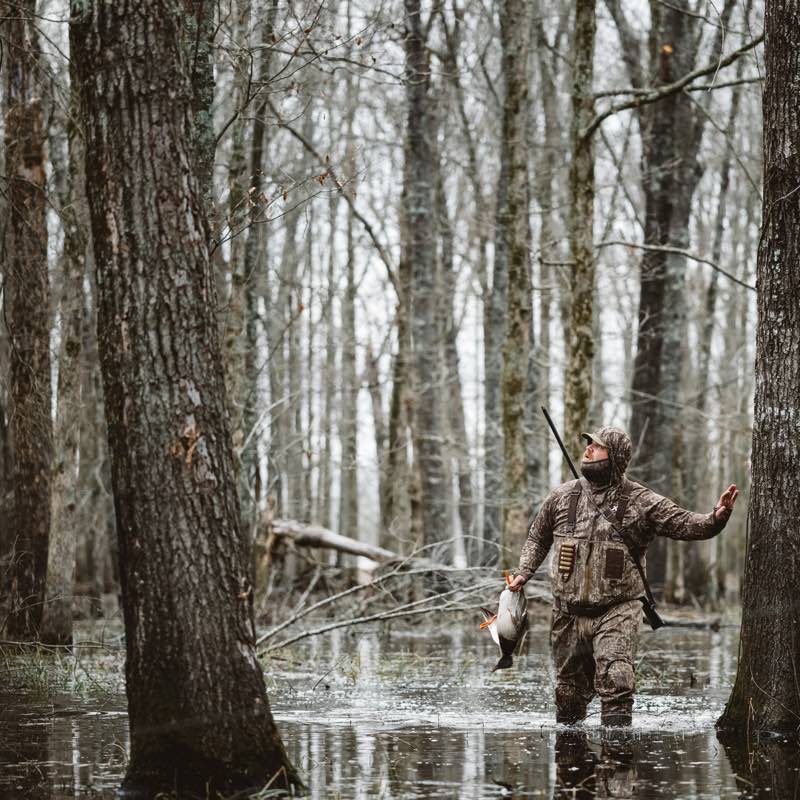 Hunter in full camouflage waders and jacket holding a duck, standing in flooded woods.