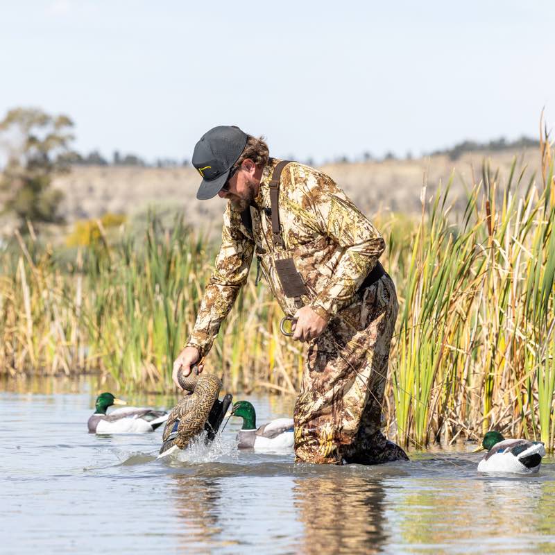 Man in camouflage hunting waders with brown suspenders adjusts duck decoy.