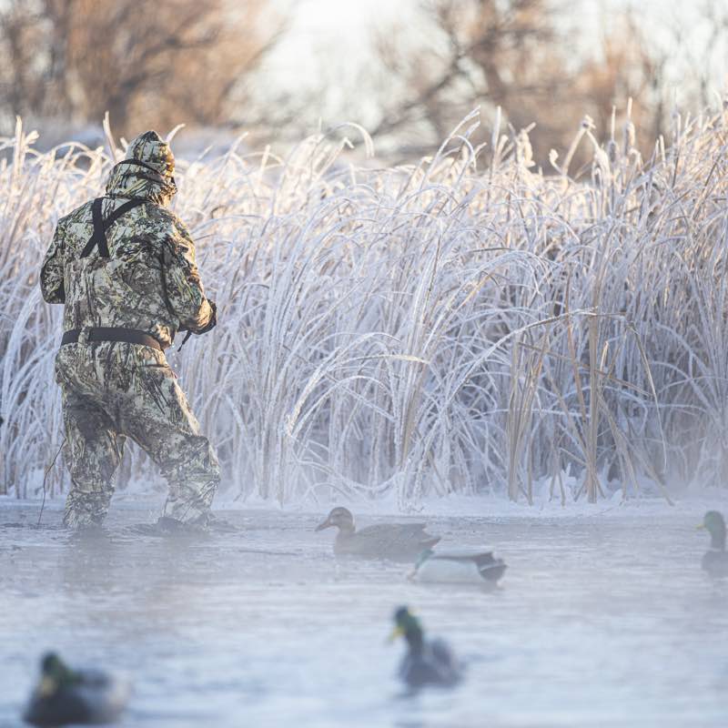 Green/brown camo hunting apparel with black 'X' back straps, wading in icy water.