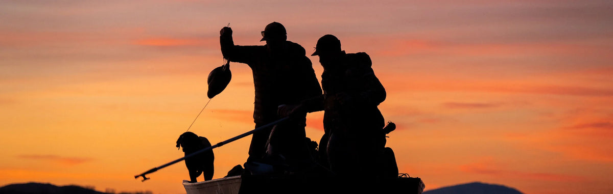 Silhouette of two hunters, dog, and decoy in boat at orange sunset.