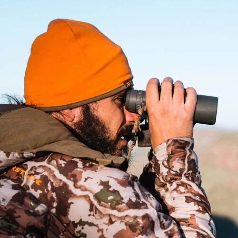 Man in orange beanie using dark binoculars with a strap.