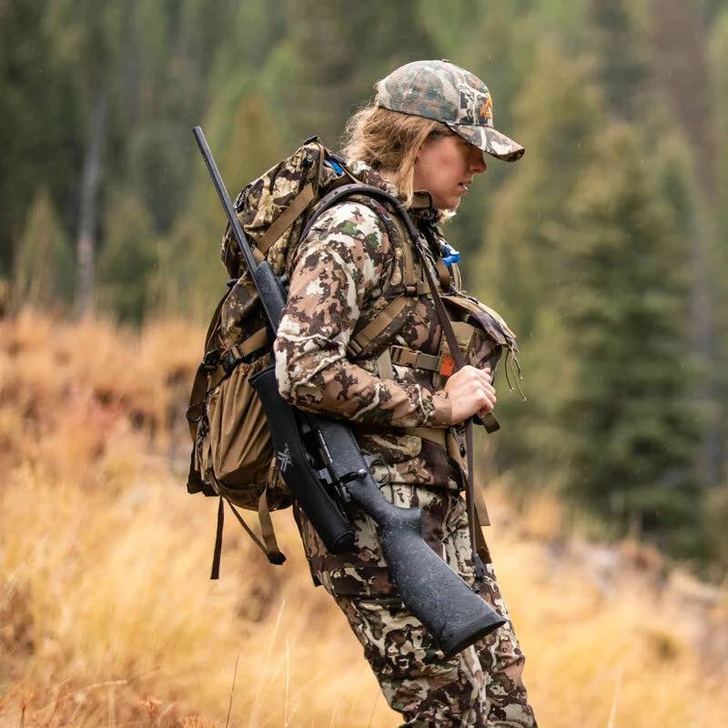 Hunter in multi-pattern camo clothing and backpack with rifle, walking outdoors.