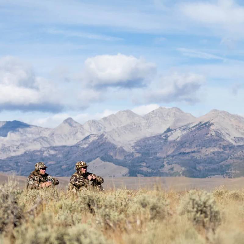 Two hunters in brown-green camouflage hunting gear with hats, sitting outdoors.
