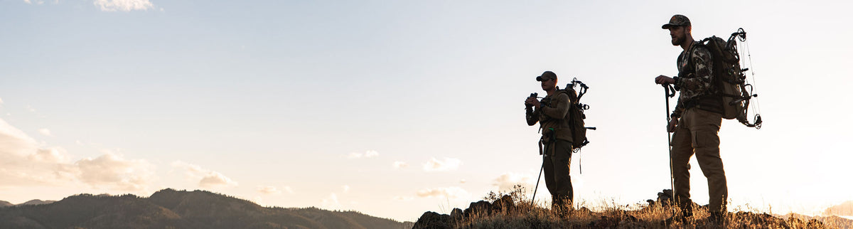 Outdoor gear: 2 men with backpacks, a bow, binoculars, and trekking pole on a ridge.
