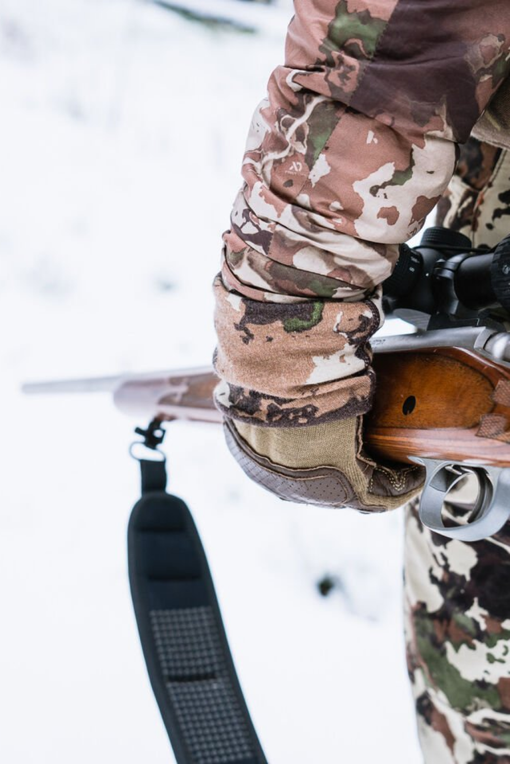 Close-up: Brown/green camo jacket & tan glove holding rifle with wooden stock and scope.
