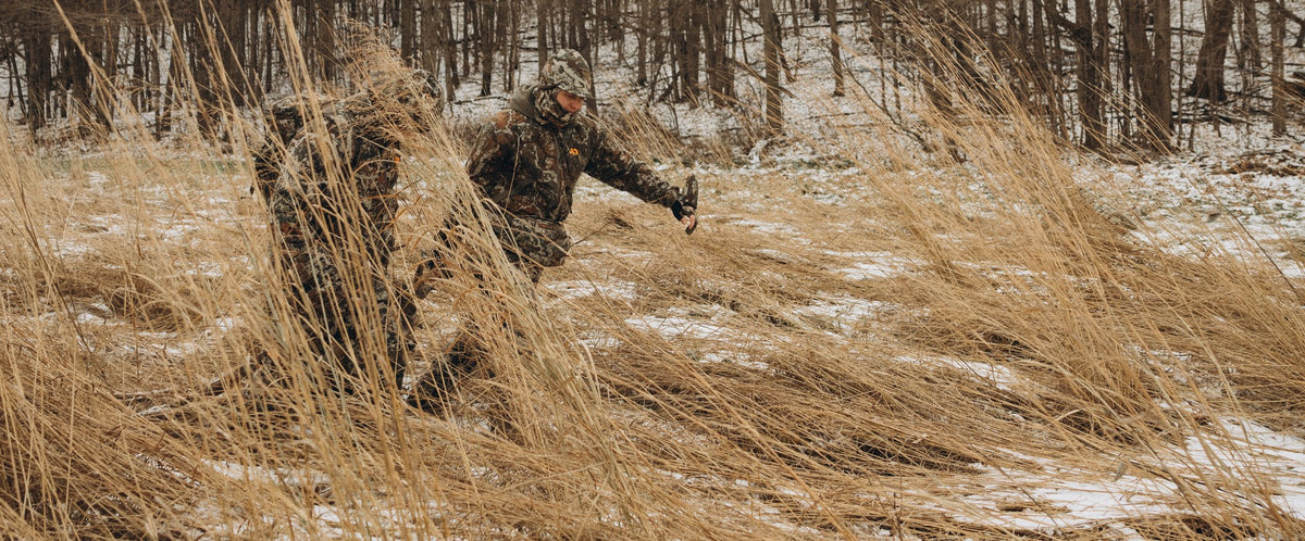 A person in a heavy camouflage hunting suit and hood crouches among tall dry grass in a snowy wooded field.