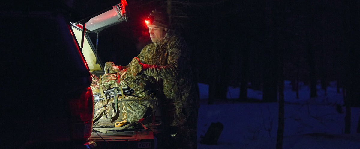 A person in camouflage clothing stands beside an open vehicle trunk at night, handling a large bundled item illuminated by a red headlamp and bright light inside the trunk, with a snowy forest in the background.