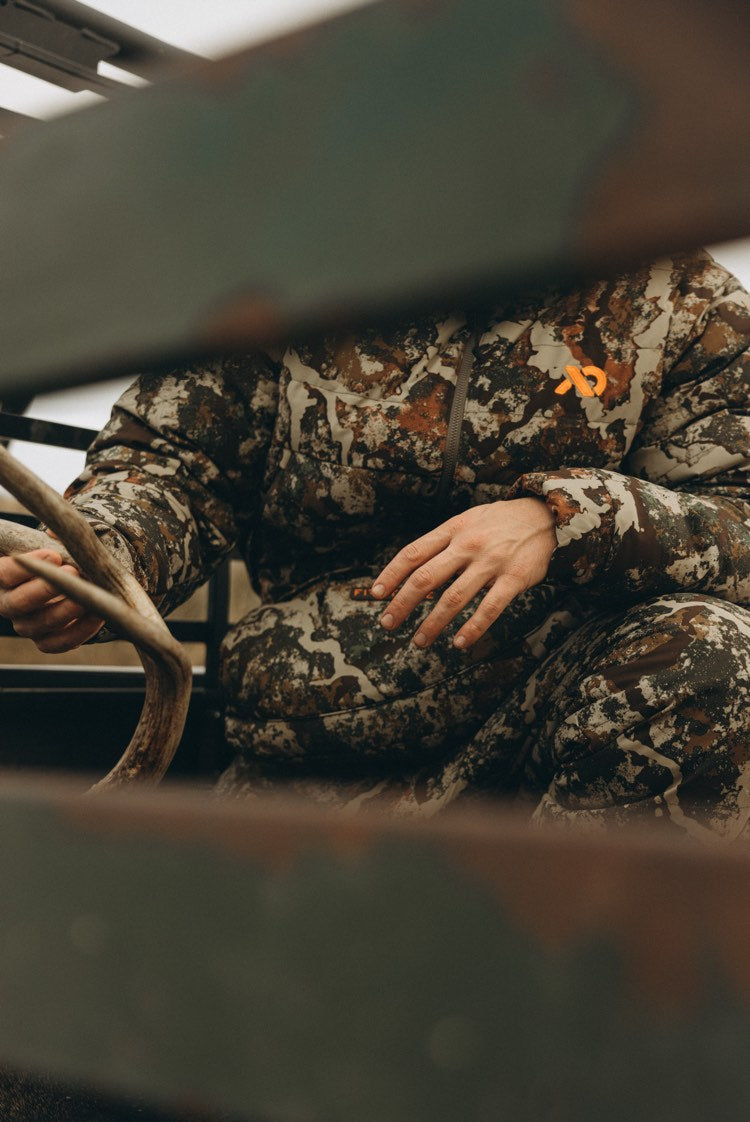 A person wearing a camouflage hunting hoodie and matching pants sits inside a vehicle, holding a deer antler. The camouflage pattern is brown, beige, and gray with a small orange logo on the chest.