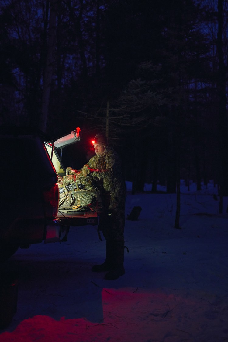 A person in camouflage stands beside an open vehicle trunk in a snowy forest at night, handling a large bundled item illuminated by red and white lights.