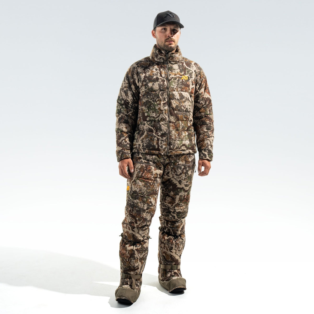 Man wearing camo jacket, pants and overboots, and a black FIRST LITE cap.