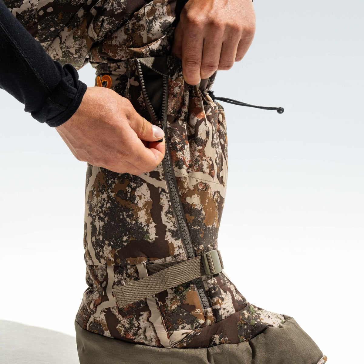 Man zipping up his camo overboot, he is wearing camo pants and black shirt.