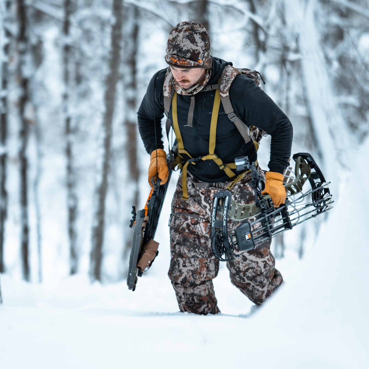 Man walking in a snowy forest carring bowas and arrows and wearing black shirt and camo hat and pants.