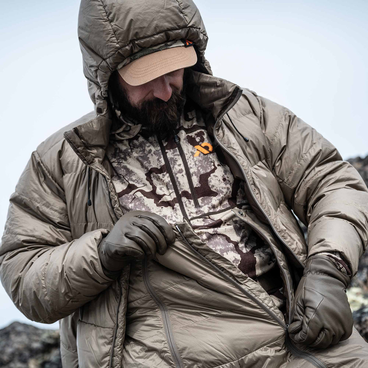 A man wearing gloves zips his Walnut hooded puffy jacket, revealing a patterned base layer shirt with an orange logo.