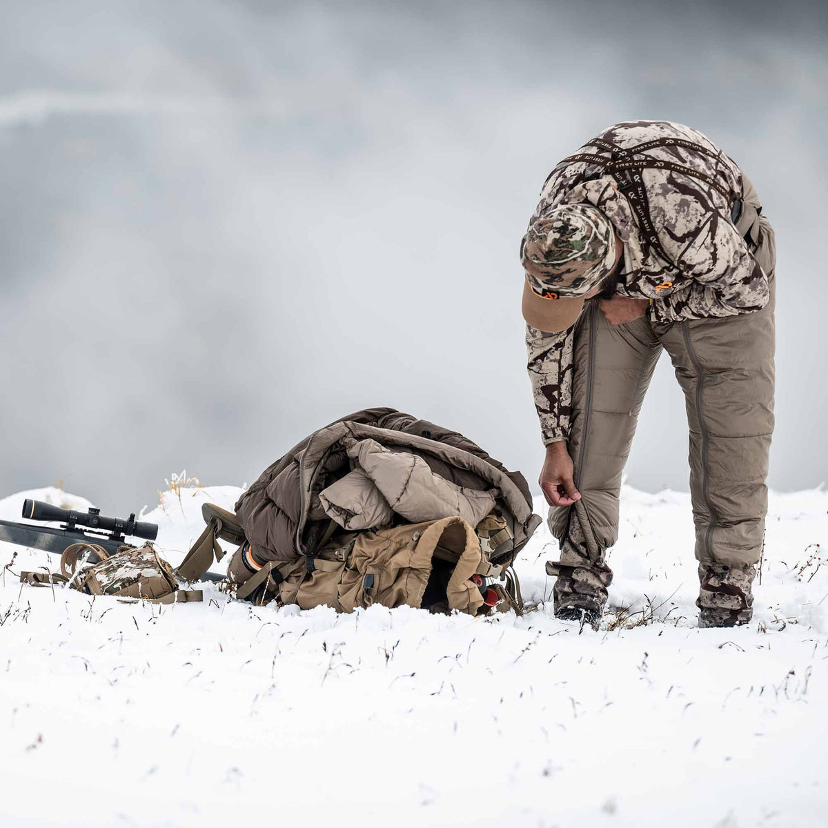 A person in camouflage adjusts light brown full-zip puffy insulated pants over their legs in a snowy landscape.