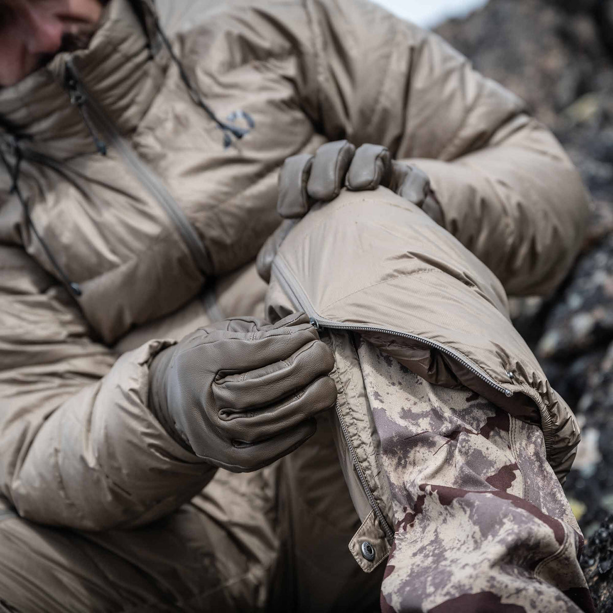 A person in tan gloves zipping a puffy tan pant leg over a camouflage inner layer.