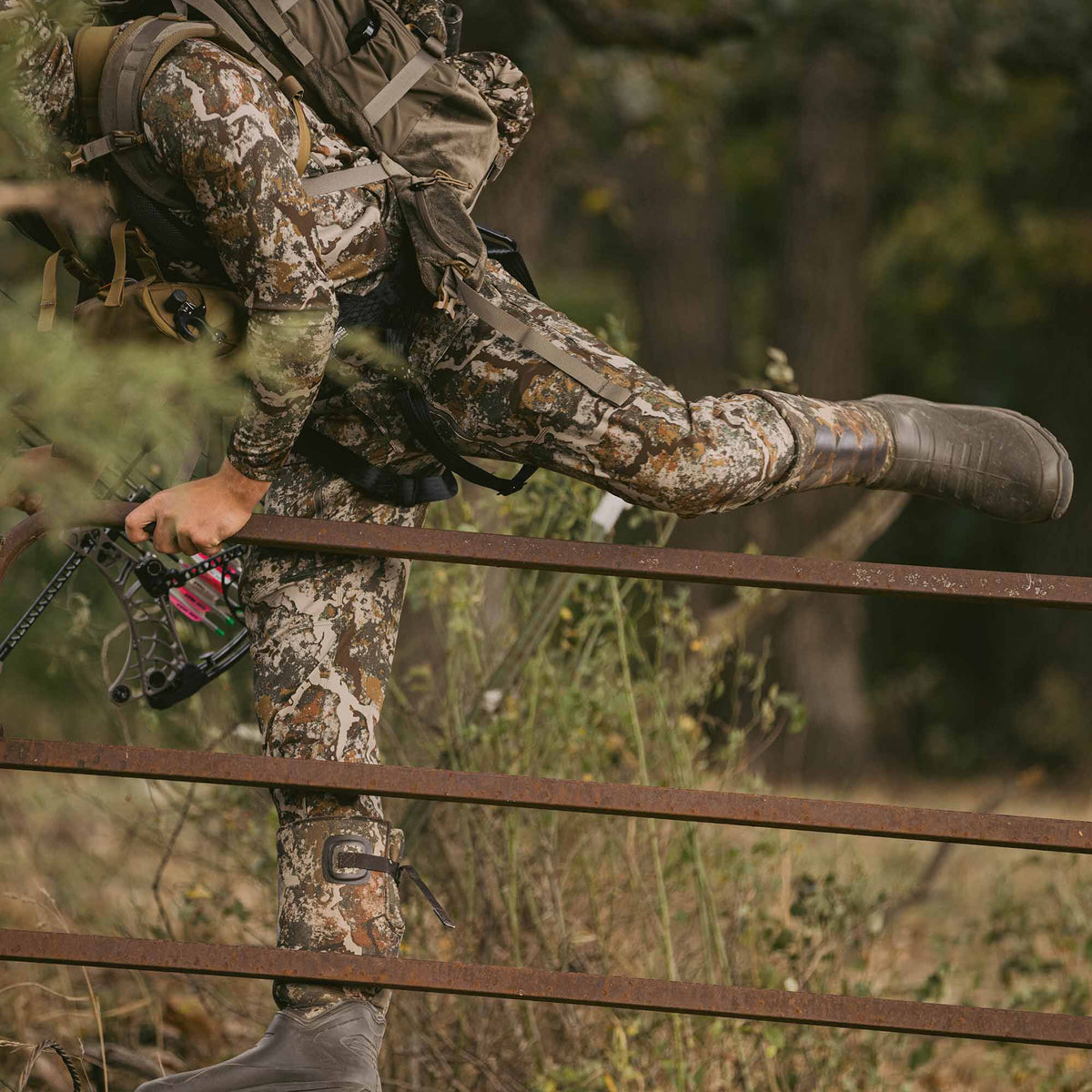 A person wearing multi-color camouflage hunting apparel and dark grey boots steps over a rusty fence, holding a bow with arrows.