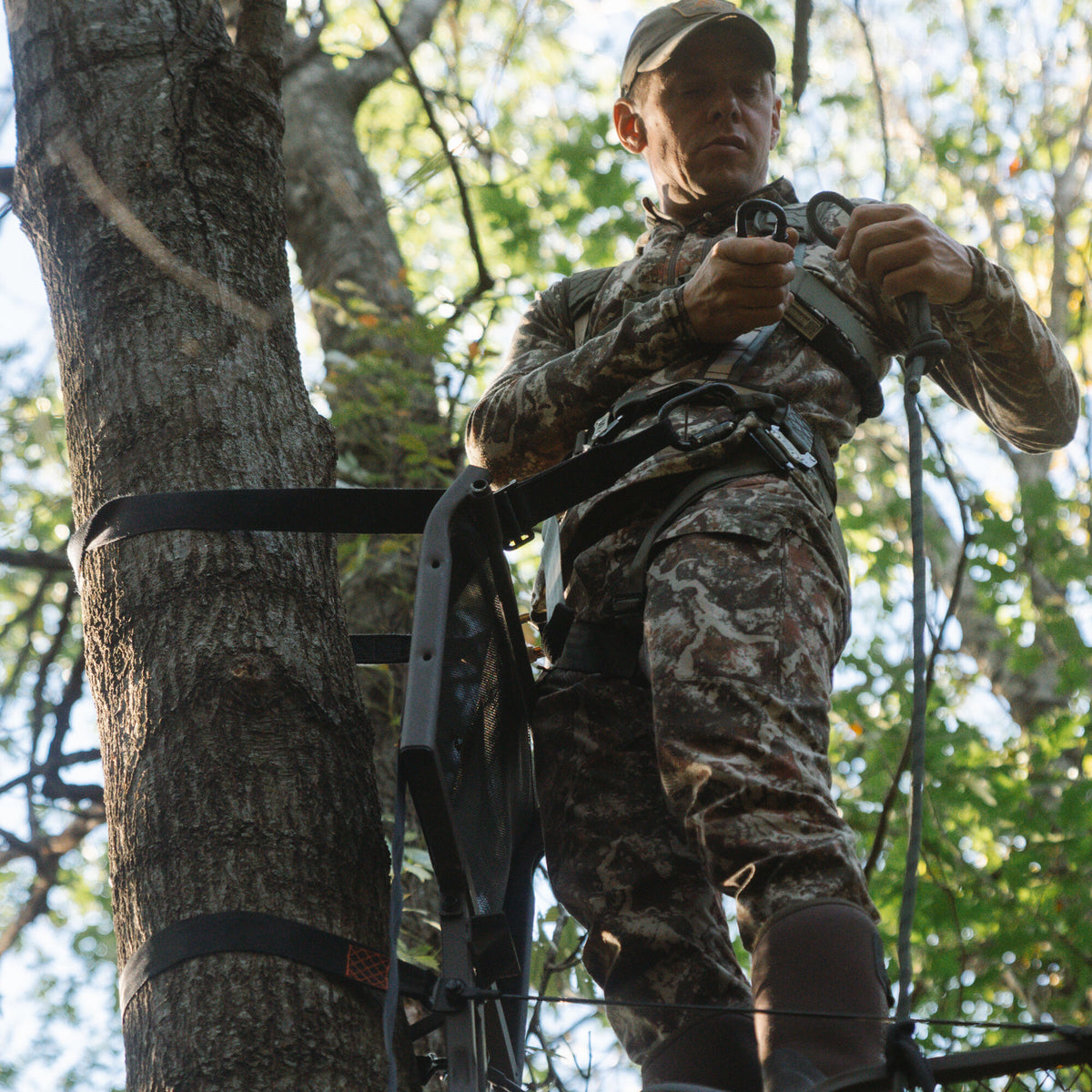 A person in camouflage uses a black tree climbing stick system with black straps and a harness to ascend a tree.