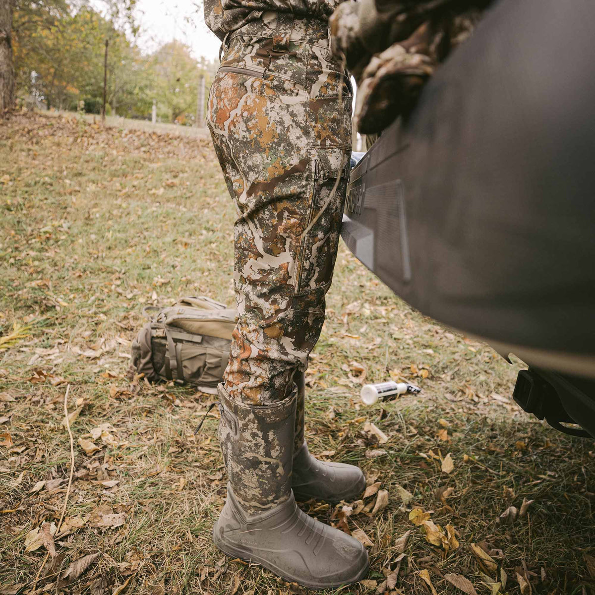 Mud-covered dark olive rubber boots and multi-color autumn camouflage pants with side cargo pockets are worn in a natural outdoor setting.
