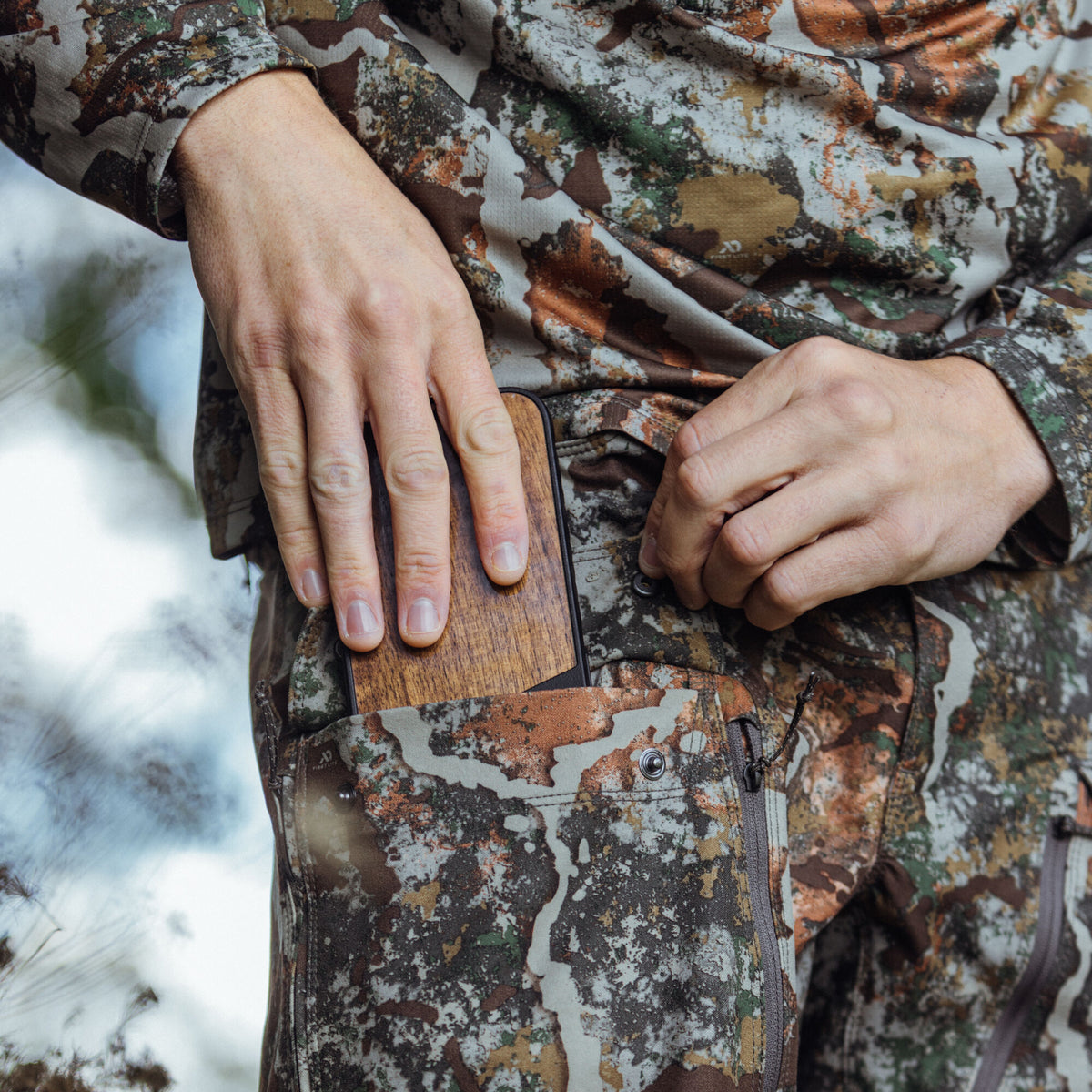 Person in First Lite multi-color camouflage gear putting a phone with a wood-grain case into a leg pocket.