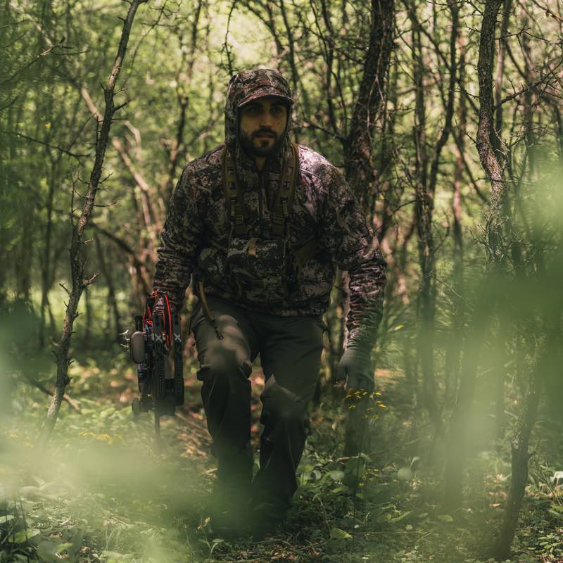 Camo hunting hoodie and cap on a man carrying a bow and chest rig in a forest.