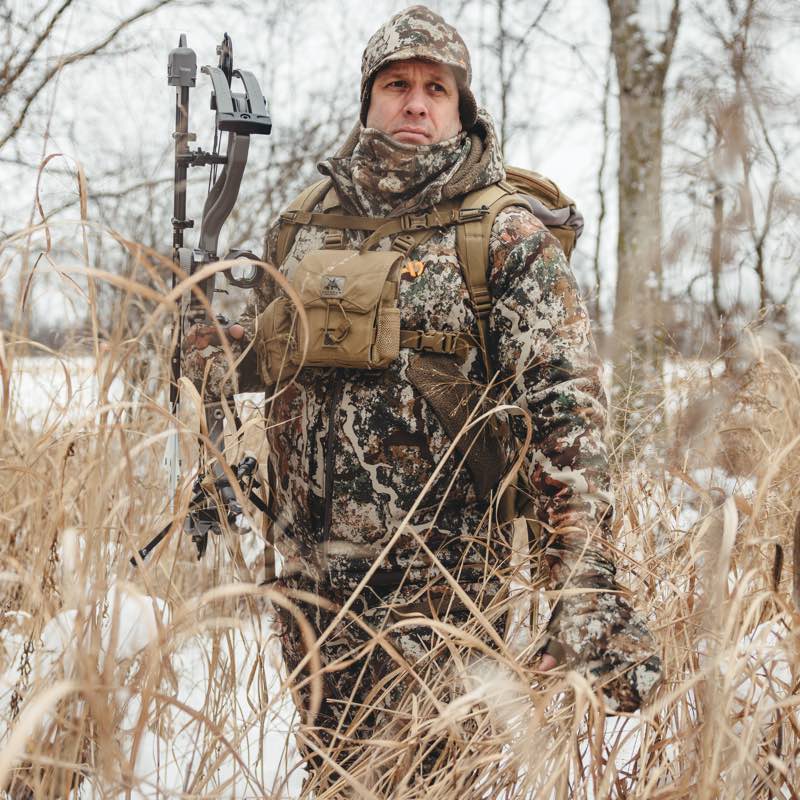 Hunter in multi-tone camo jacket, hat, and chest pack, holding a compound bow.