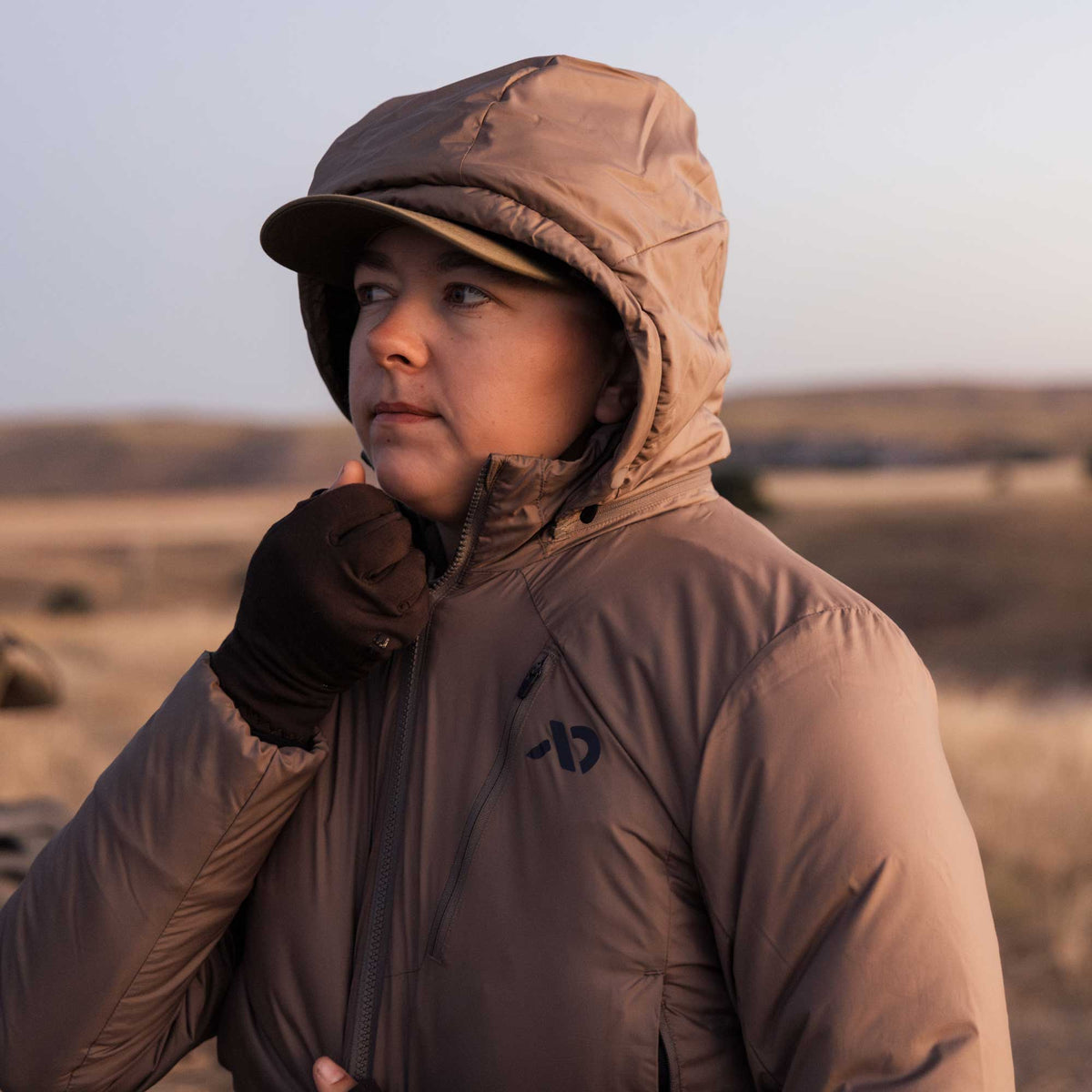 Tan hooded insulated jacket with a front zipper, a zippered chest pocket, and a dark brand logo.