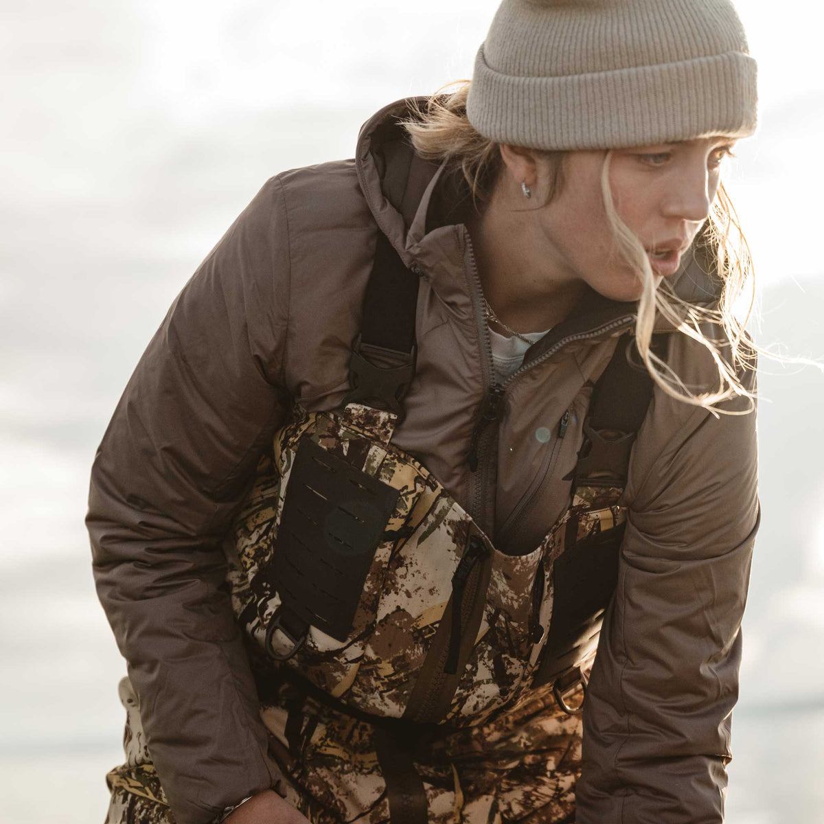 Brown hooded puffer jacket worn over camouflage bib waders with a visible chest pocket.