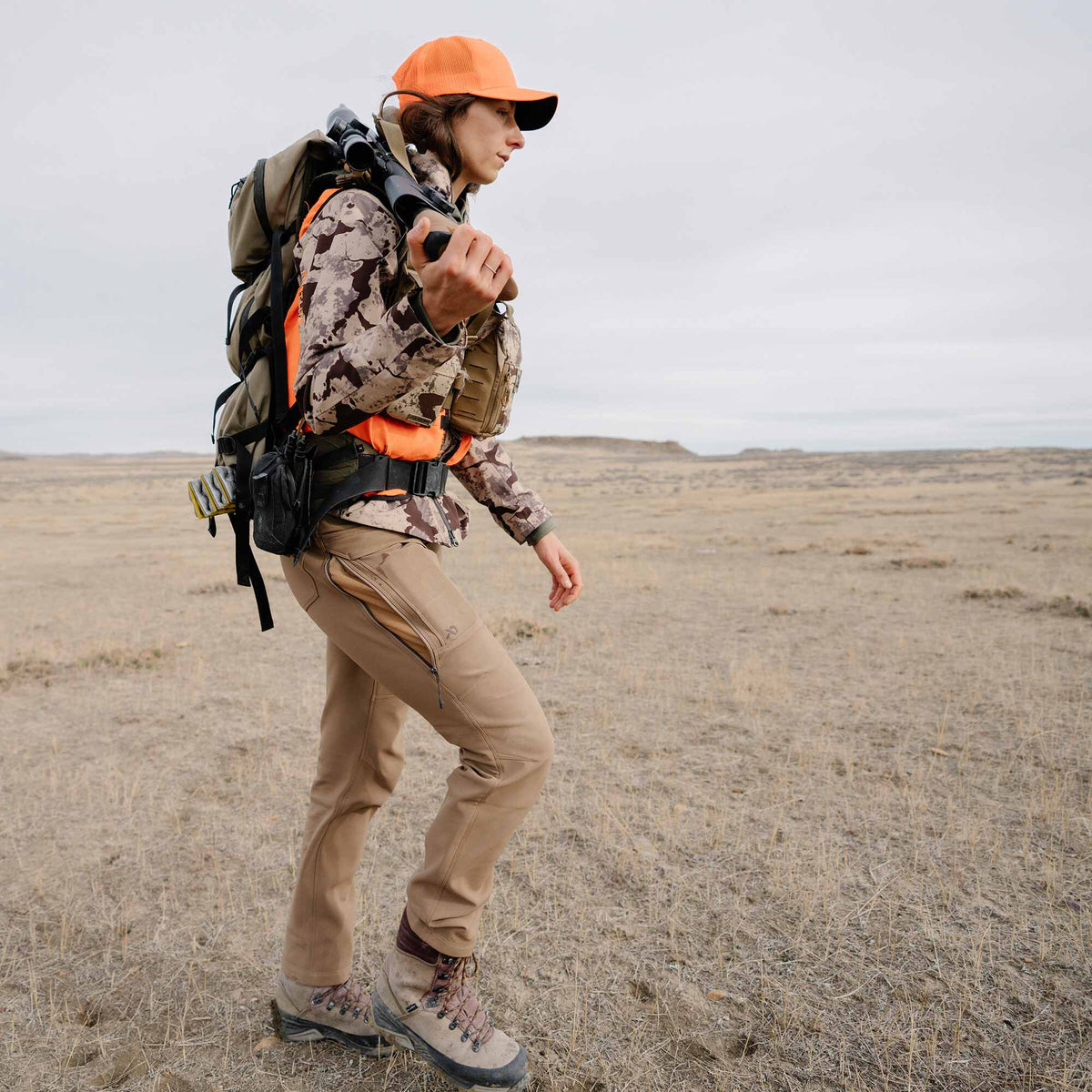 Tan outdoor pants featuring side leg zippers with mesh ventilation, modeled by a person walking in a dry field.