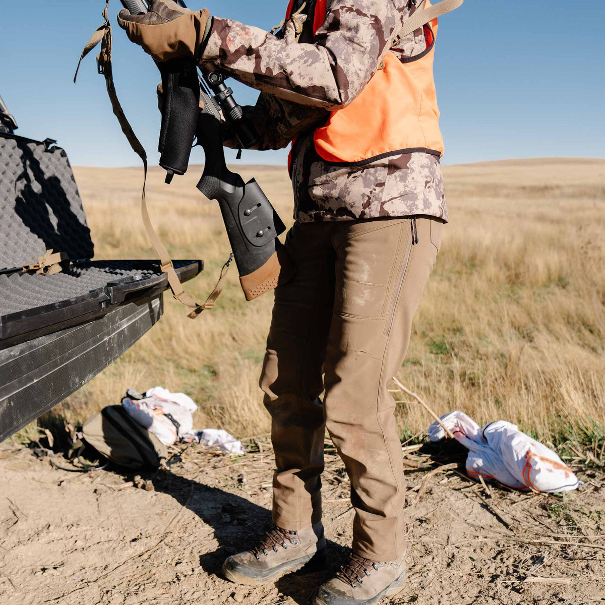 Hunter wearing a camouflage jacket, orange vest, and tan tactical pants holds a rifle with a scope.