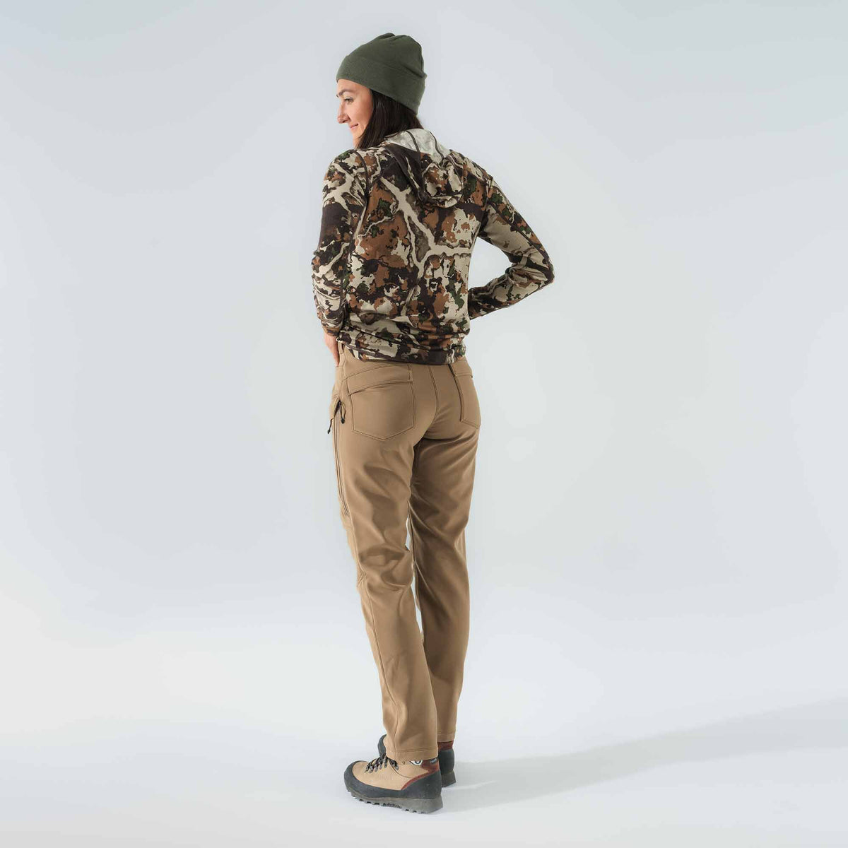 Woman wearing a camouflage hooded long-sleeve shirt, tan hiking pants with a zippered side pocket, an olive green beanie, and tan hiking boots.
