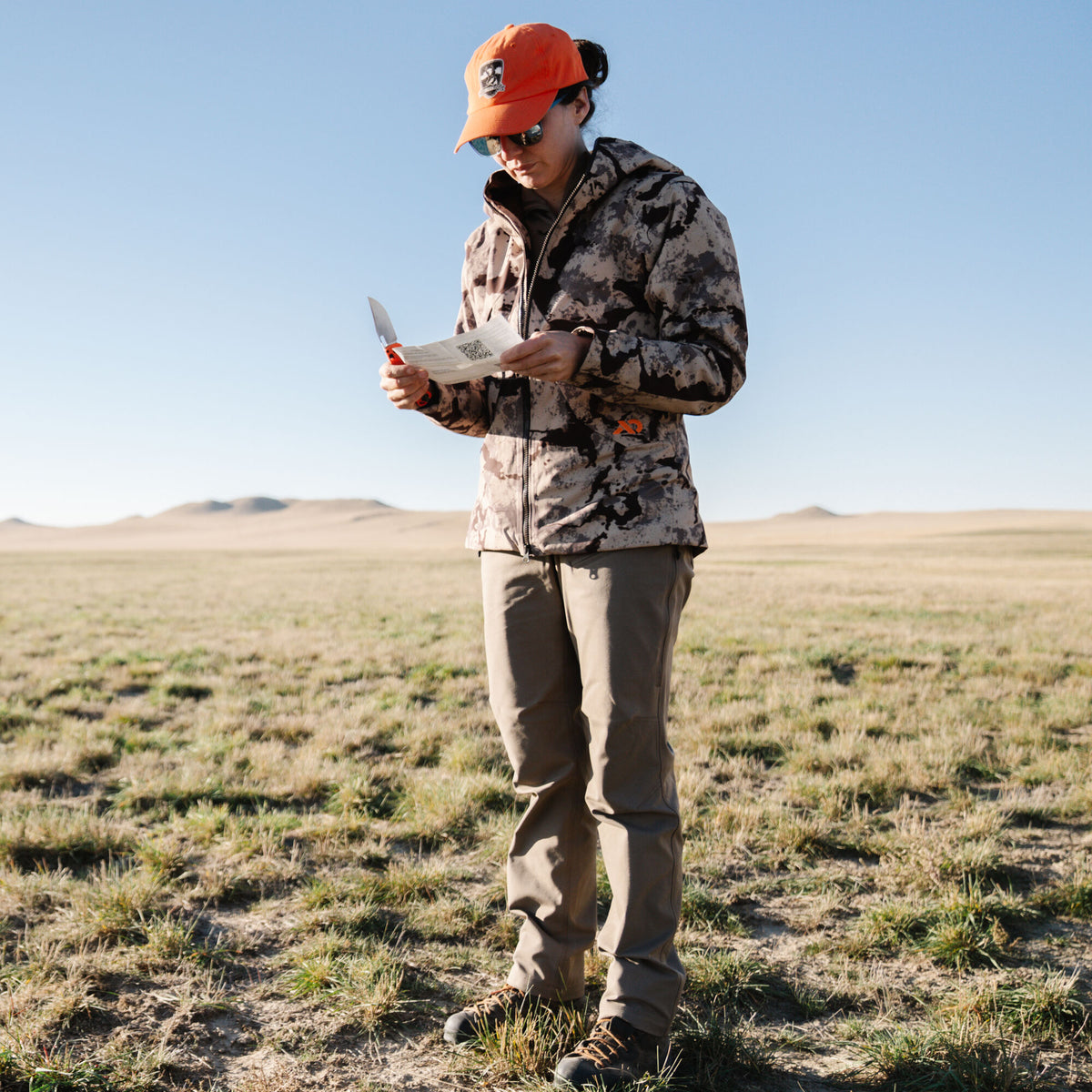 Person in a tan camouflage jacket, brown pants, and orange hat holds an orange-handled knife and paper.