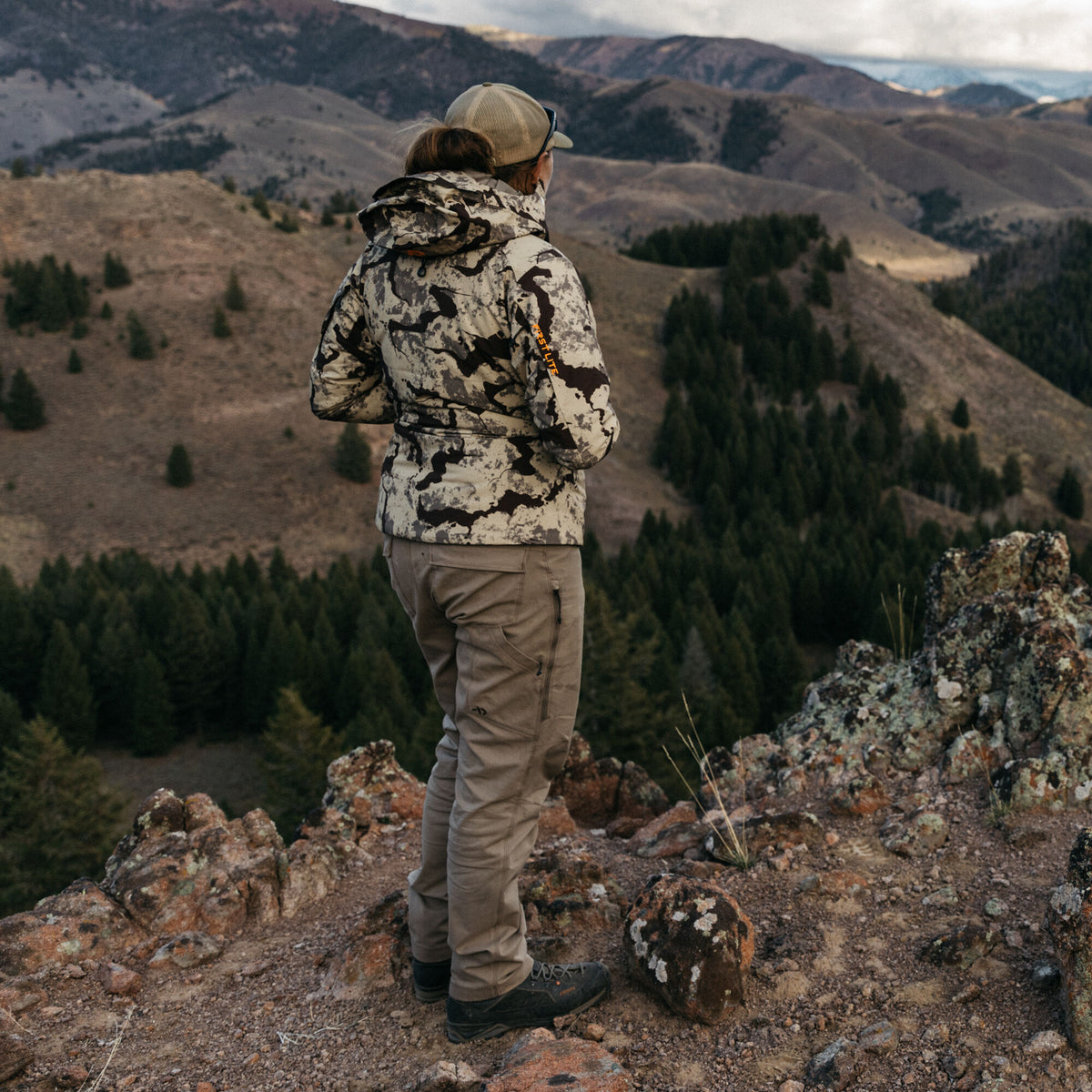 A person wears a tan and dark brown camouflage First Lite hunting jacket with a hood and tan cargo pants, standing on a mountain overlooking a valley.