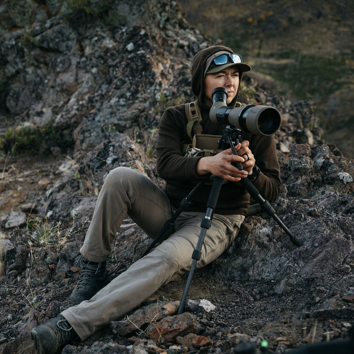 A person in a dark brown hoodie and olive-tan pants sits on a rocky slope, looking through a tan and black Sig Sauer angled spotting scope on a tripod.