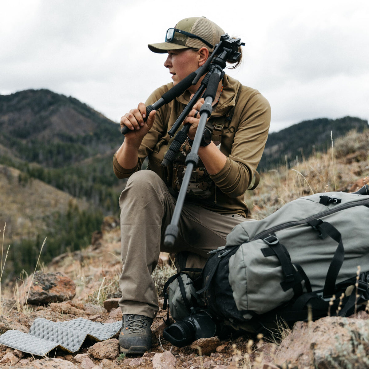 A person in outdoor clothing adjusts a black tripod on rocky terrain.