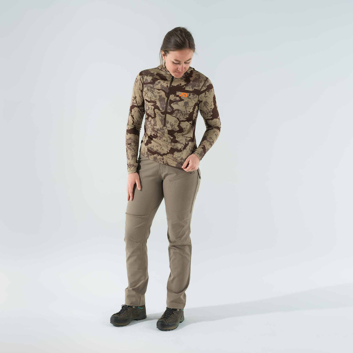 A woman wears a brown camouflage long-sleeve quarter-zip top with an orange logo and khaki outdoor pants with a zippered thigh pocket.