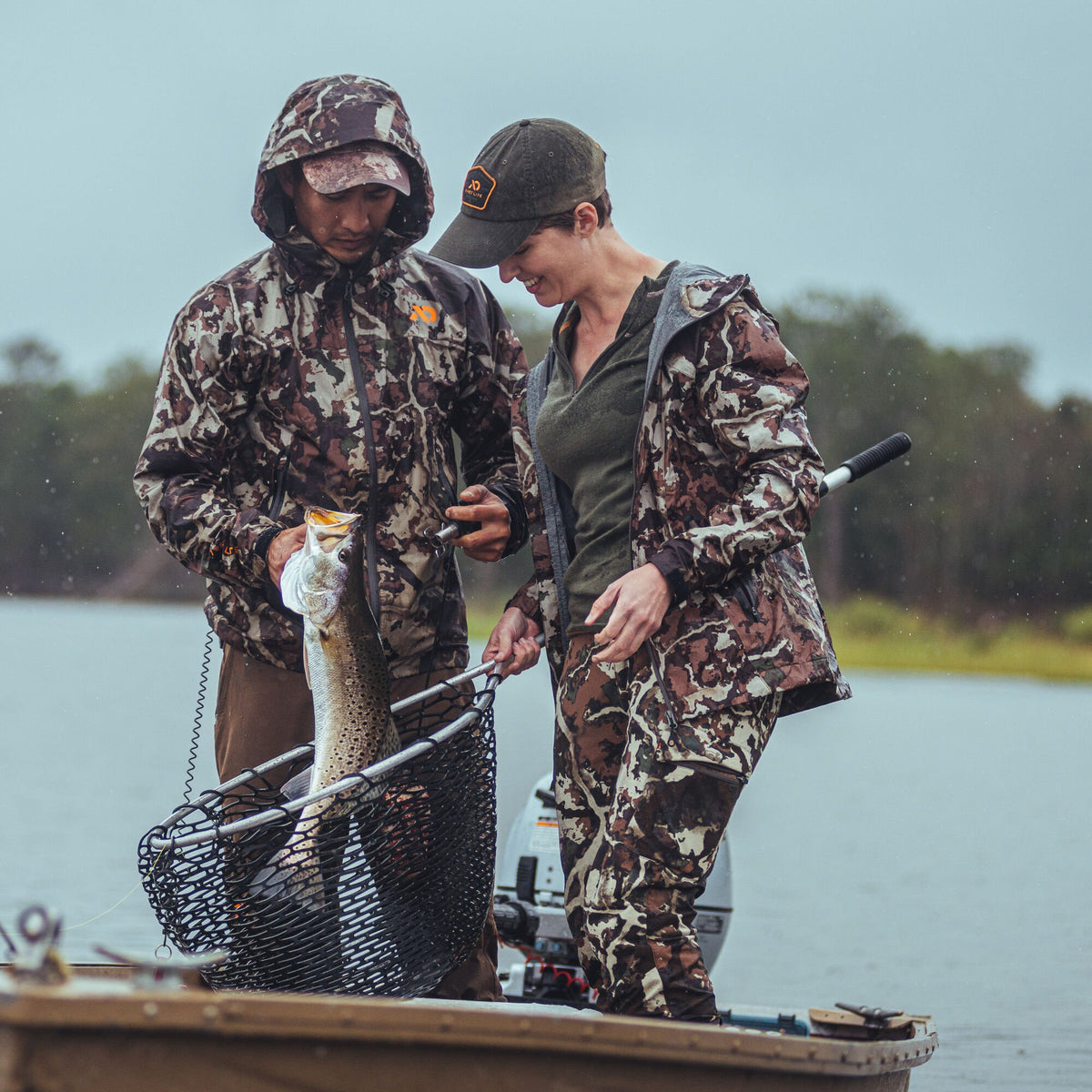 Two anglers in First Lite Fusion camouflage hooded jackets and matching pants on a boat, holding a speckled trout in a net.